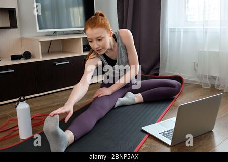 Das rothaarige Mädchen ist in einem Haus Stretching sitzt auf einer Sportmatte vor einem Laptop engagiert Stockfoto