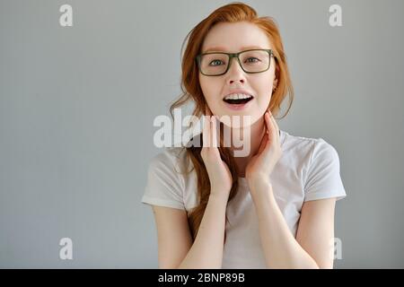 Ein rothaariges Mädchen mit Brille steht auf einem grauen Hintergrund und schaut glücklich in die Kamera und glättet ihr Haar. Stockfoto