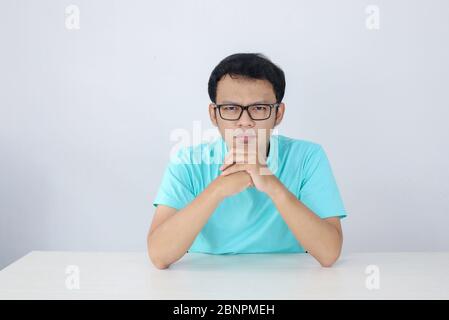 Junge asiatische Mann tragen blaues Hemd und Brille mit ernsten Gesicht auf die Kamera schauen und Hand halten Kinn auf dem Tisch. Stockfoto