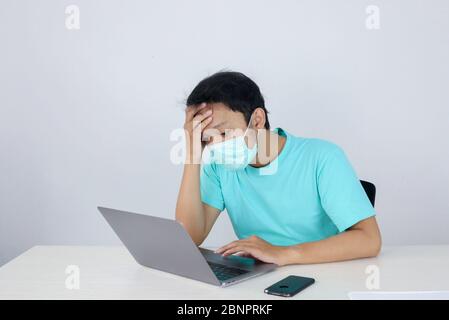 Junge asiatische Mann trägt medizinische Maske fühlt sich ungesund, müde, und verwirrt mit der Arbeit in Laptops auf dem Tisch. Stockfoto