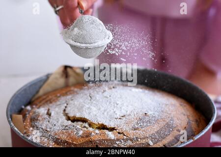 Frau Hand streuen Puderzucker über frischen Biskuitkuchen. Puderzucker fällt auf frischen perfekten Marmorkuchen/ Rezepte und Ideen für Frühstückskuchen Stockfoto