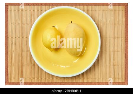 Gelbe Früchte - helle Zitrone und helle Mango - auf einem gelben Teller auf einer Zuckerrohrtischdecke Stockfoto