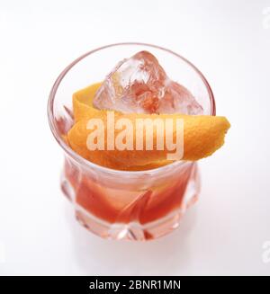 Rötlich-brauner Cocktail in einem Felsglas auf einem isolierten weißen Hintergrund. Der Cocktail wird mit einem orangen Twist und handgeschnittenem Eis garniert. Stockfoto