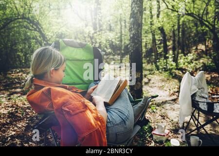Finden Einsamkeit in Wildnis Konzept. Junge Frau, die im Wald ruht, auf dem Lagerstuhl sitzt und Buch liest Stockfoto