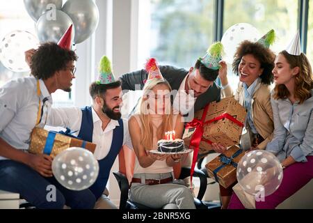 Geschäftsfrau bläst Kerzen auf ihrem Geburtstagskuchen surpise Geburtstagsfeier im Büro. Mitarbeiterfeier mit Dekorationen. Stockfoto