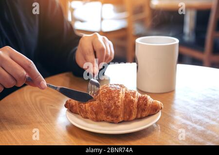 Nahaufnahme Bild von Händen Schneiden ein Stück Croissant mit Messer und Gabel zum Frühstück mit Kaffeetasse auf Holztisch Stockfoto