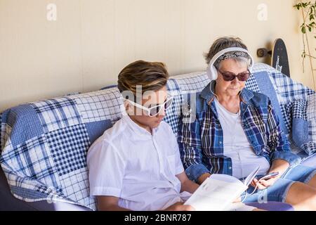 Ein Paar Mann und Frau sitzen auf dem Sofa - zwei verschiedene Generationen zusammen ein Buch lesen und Musik hören mit Kopfhörern des Telefons - Senior und Teenager Stockfoto