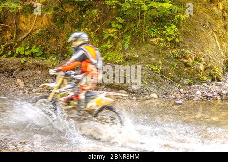 Sonniger Sommertag. Enduro-Athlet überwindet einen Waldbach. Sonnenlicht und viel Wasser Stockfoto