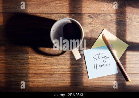 Kaffeetasse, Bleistift und Notizpapier mit Stay Home-Nachricht auf Holzschreibtisch mit Fenstersonnenlichteinfall. Quarantäne oder Isolierung des Coronavirus. Stockfoto