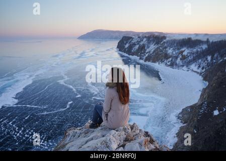 Baikalsee im Winter. Frau sitzt auf einer Klippe und schaut auf den gefrorenen Baikalsee. Tiefster und größter Süßwassersee. Olchon, Russland, Sibirien Stockfoto