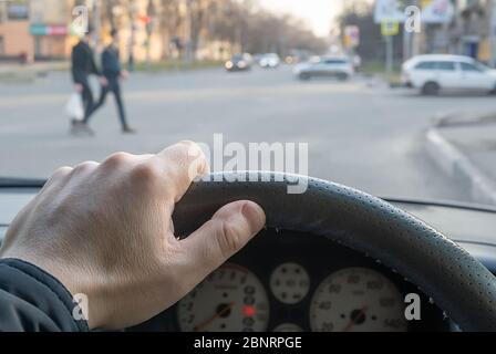 Blick aus dem Auto, Hand des Mannes am Lenkrad des Autos, befindet sich gegenüber der Fußgängerampel und Fußgänger über die Straße Stockfoto