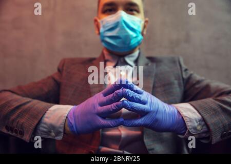 Der junge elegante Mann sitzt in einem Ledersessel mit Maske im Gesicht und medizinischen Handschuhen auf Betongrund. Nahaufnahme der Hände. Stockfoto