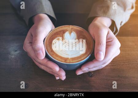 Nahaufnahme Bild der Hände halten eine blaue Tasse heißen Latte Kaffee mit Latte Kunst auf Holztisch im Café Stockfoto