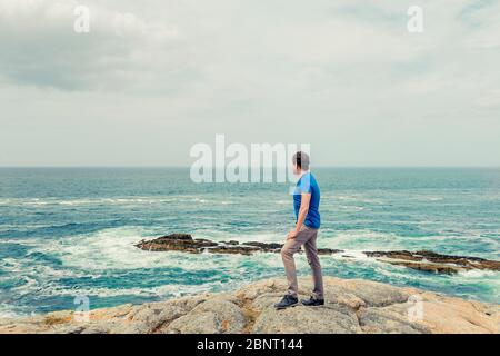 Mann in blauem Hemd, der auf den Felsen steht und das raue Meer beobachtet Stockfoto