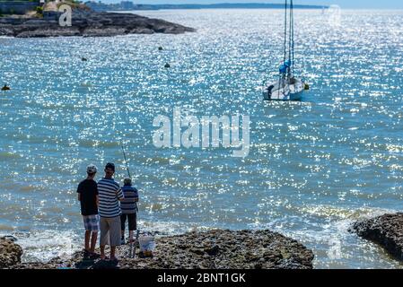 Saint-Palais-sur-Mer, Frankreich: Drei Männer fischen auf den Felsen in der Conche de Saint Palais mit einem Segelboot und der Corniche de Nauzan dahinter. Stockfoto