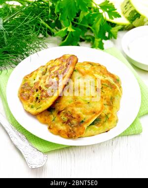 Fritten von Zucchini, Dill und Petersilie in einem Teller auf einem Handtuch, saure Sahne in Untertasse auf dem Hintergrund von hellen Holzbrett Stockfoto