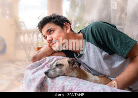 Der junge Mann umarmt einen Hund, der auf einem Sessel im Garten liegt, und lächelt Stockfoto