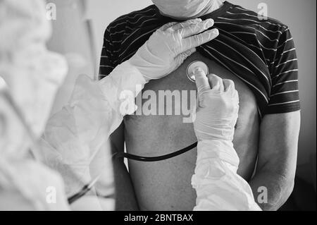 Zugeschnittene Nahaufnahme eines Arztes in einem Schutzanzug, der ältere männliche Patienten zu Hause mit Phonendoskop untersucht, Brustkontrolle, Schwarz-Weiß, COVID-19-Konzept Stockfoto