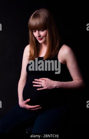 Porträt einer nachdenklichen schwangeren Frau mit schwarzem Hintergrund, nach unten schauend. Stockfoto