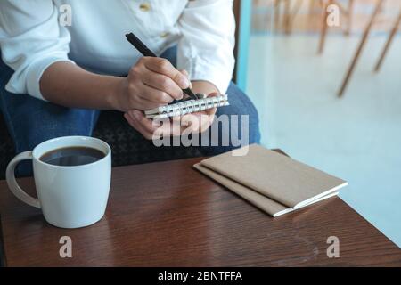 Nahaufnahme Bild einer Frau Hände halten und schreiben auf leere Notizbuch mit Kaffeetasse auf Holztisch Stockfoto