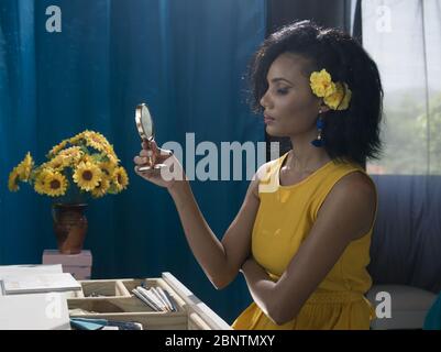 25-30 junge Brünette Mädchen an Eitelkeit tragen lange gelbe Kleid mit Sonnenblumen und blauen Vorhang ne im Hintergrund , Stockfoto