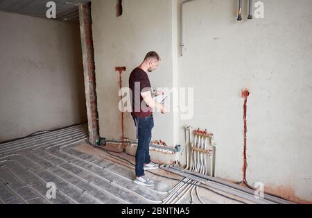 Junger Mann in leerer unvollendeter Wohnung, die Wand mit Hilfe von Maßband vermessen, Notizen überprüfen, auf Fußbodenheizung Rohrsystem stehen. Konzept der Reparatur und Sanierung. Stockfoto