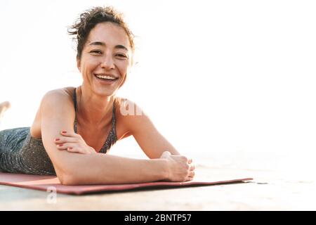 Foto von fröhlichen Sportlerin im Trainingsanzug lächelnd und liegen auf Fitness Matte beim Üben von Yoga am Morgen am Meer Stockfoto