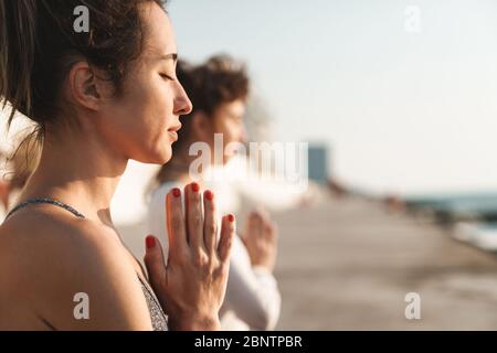 Foto von motiviert junge Frauen in Trainingsanzügen meditieren und die Handflächen zusammen beim Üben von Yoga am Morgen im Freien Stockfoto