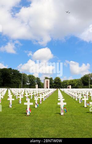 Grabsteine mit Fahnen und Blumen auf dem Luxembourg American Cemetery und Memorial Memorial Day Wochenende. Ein Flugzeug fliegt durch die Wolken. Stockfoto