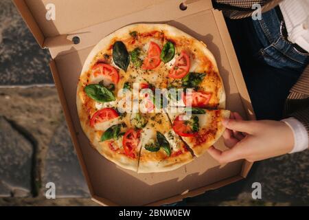Draufsicht Bild von paar Schnapp Scheiben Pizza aus der Box im Freien. Mann und Frau Hände, die Pizza nehmen. Vegane Pizza mit frischen Tomaten Basilikum und Stockfoto