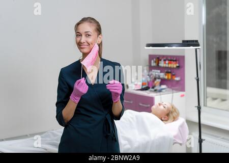 Kosmetologie Kabinett Client liegt auf Couch Kosmetiker steht rosa schützende medizinische Maske Handschuh und Lächeln Vorbereitung für das Verfahren permanente Augentrost Stockfoto