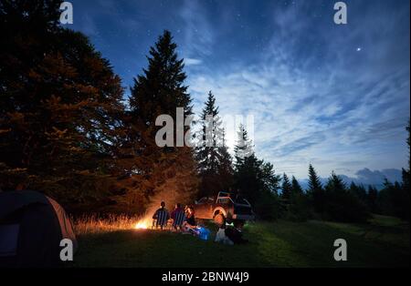 Gruppe von Reisenden, die in der Nähe von Lagerfeuer im Wald mit hohen Nadelbäumen, Auto und Zelt unter dem schönen Nachthimmel sitzen. Fantastische Aussicht auf blauen Sternenhimmel über Wiese mit Reisenden in der Nähe Lagerfeuer. Stockfoto