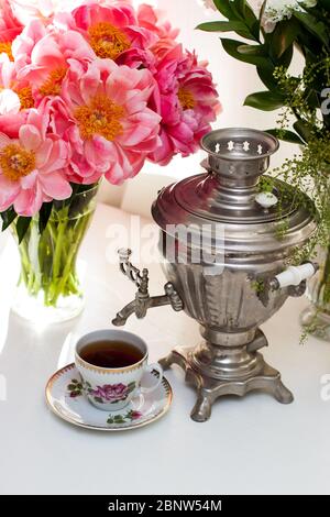 Stillleben mit altem russischen Samowar, rustikaler Tasse Tee, Bouquet mit Pfingstrosen, Gläsern und Kerzen. Gemütliche Teezeremonie in weiß gefärbtem Interieur. Stockfoto