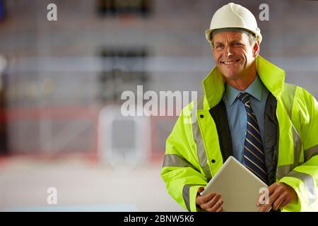 Porträt eines lächelnden Architekten, der einen Hardhat und reflektierende Kleidung trägt, die ein digitales Tablet auf der Baustelle hält Stockfoto