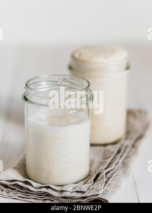 Zwei Glasgläser mit Weizensauerteig-Vorspeisen in verschiedenen Flüssigkeitsstufen. Starter 100 Prozent Hydratation im Hintergrund und Starter mit höherer Hydratation im Vordergrund. Kopierbereich. Vertikal. Stockfoto