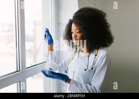 Afroamerikanische Ärztin in einem weißen Mantel mit Stethoskop. Sitzt am Fenster mit Reagenzflaschen auf weißem Hintergrund. Forscher Stockfoto