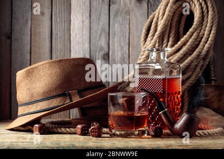 Starkes alkoholisches Getränk, Pfeife und Würfel auf einem alten Holztisch. Im Hintergrund ein Hanfseil und ein Männerhut. Konzept für die großen Jungs. Stockfoto