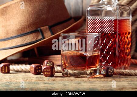 Starkes alkoholisches Getränk und Würfel auf einem alten Holztisch. Im Hintergrund ein Hanfseil und ein Männerhut. Stockfoto