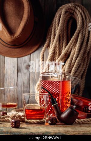 Starkes alkoholisches Getränk, Pfeife und Würfel auf einem alten Holztisch. Im Hintergrund ein Hanfseil und ein Männerhut. Konzept für die großen Jungs. Stockfoto