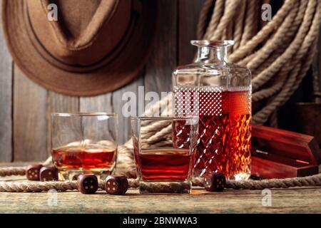 Starkes alkoholisches Getränk und Würfel auf einem alten Holztisch. Im Hintergrund ein Hanfseil und ein Männerhut. Stockfoto