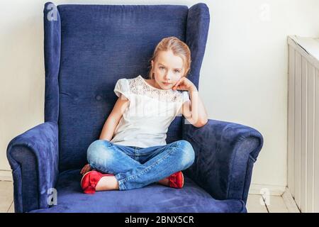 Süße kleine Mädchen in Jeans und weißen T-Shirt auf modernen gemütlichen blauen Stuhl sitzen entspannen in weißen hellen Wohnzimmer zu Hause drinnen. Kindheit Schüler Jugend entspannen Konzept Stockfoto