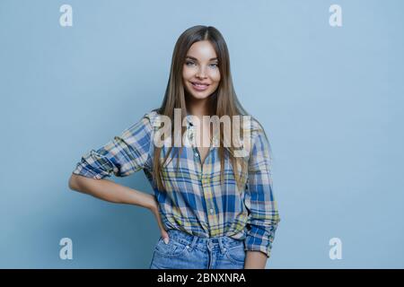 Charmante hübsche Millennial Dame hält Hand an Figur, steht selbstbewusst und lächelt fröhlich, trägt kariertes Hemd und Jeans steht in lässiger Haltung Stockfoto