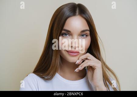 Charming gut aussehende Millennial Frau hält Hand in der Nähe Gesicht, hat gesunde leuchtende Haut, dunkles Haar, blaue Augen, in Freizeitkleidung gekleidet, isoliert über bro Stockfoto