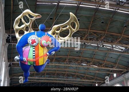 Eine Statue eines Schutzengels mit goldenen Flügeln im Zürcher Hauptbahnhof in Aufwärtsperspektive. Der Engel selbst ist sehr bunt. Stockfoto