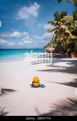 Gelbe Rucksack und Flip Flops auf schönen tropischen Sandstrand. Palmen und blauer Himmel. Urlaubskonzept Stockfoto