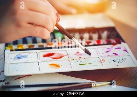 Der Künstler zeichnet mit einem Pinsel und Aquarellen in verschiedenen Farben, auf einer Kunststoffpalette, und daneben liegt ein Blatt Papier und andere Pinsel, Stockfoto