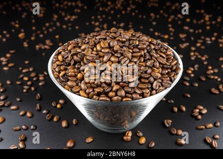 Frische, geröstete Kaffeebohnen in einer Glasschüssel, die auf einem schwarzen Tisch stehen, mit viel Kaffee verstreut. Stockfoto