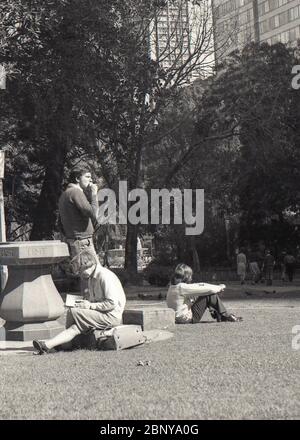 Sydney Australien 1977: Mittagspause im Hyde Park in Sydney liest eine Frau in der warmen Sonne und ein anderer Mann sitzt einfach auf dem Gras, während eine dritte Person steht und eine Zigarette raucht Stockfoto