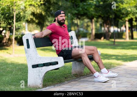 Porträt eines glücklichen jungen Mannes, der sich in einer Parkbank ausruht, nachdem er im Freien joggt Stockfoto