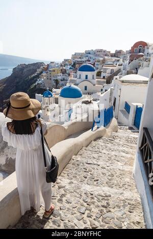 Oia, Santorini, Griechenland - 22. Juli 2014: Weibliche Touristen mit weißem Kleid und Strohhut, die das griechische Dorf mit der blauen Kuppelkirche sehen Stockfoto